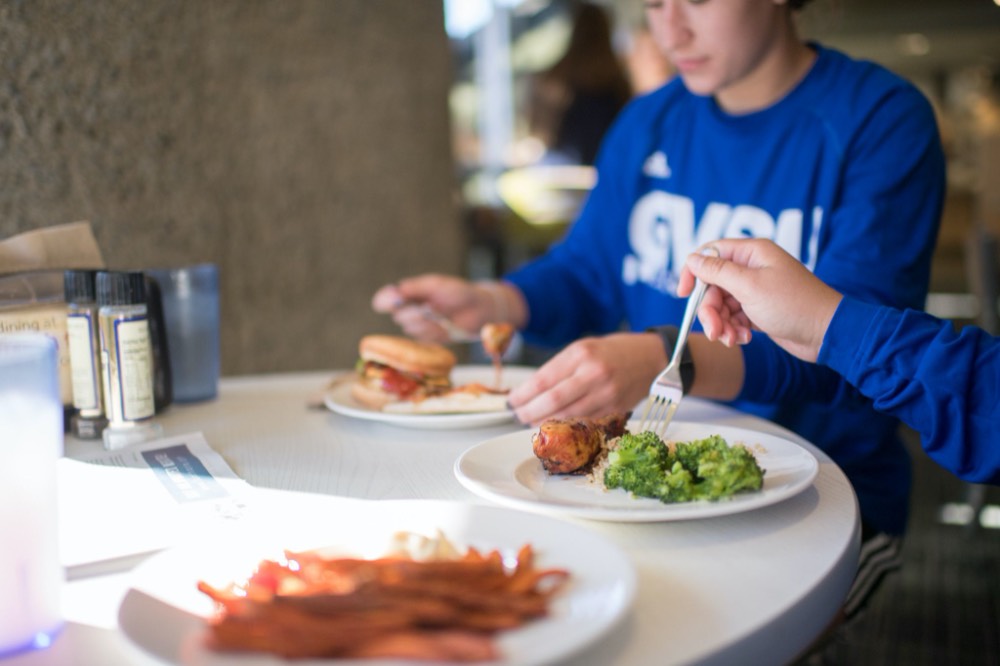 Three plates of food, one with broccoli and a chicken wing, and a hamburger. Two students are seen in the photo.